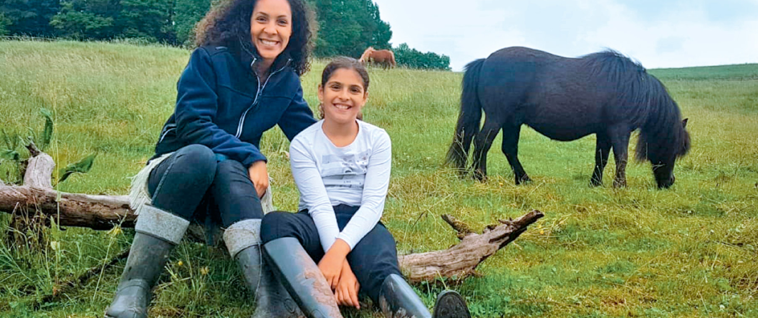 Mutter und Tochter mit Pony im Hintergrund auf einer Wiese als Startbild der Seite Pferdegestütztes Coaching und Workshops