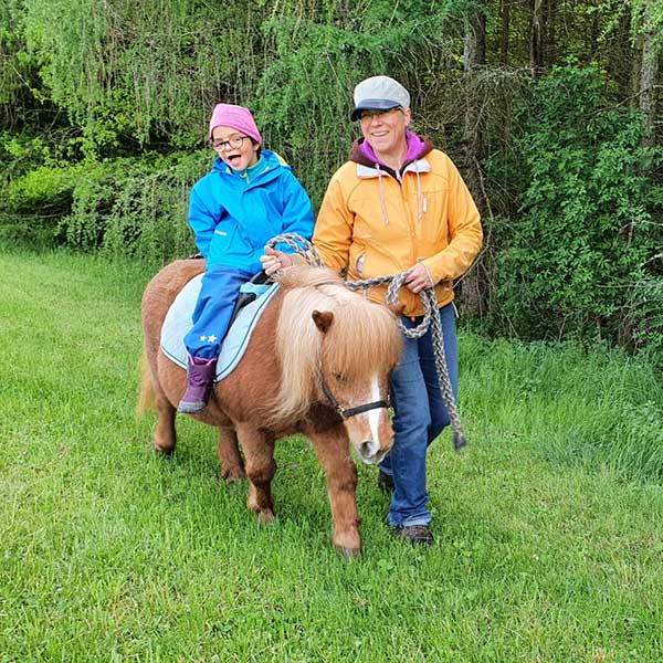 Shetlandstute Whisky mit Kind auf dem Rücken wird von Karin Miller über eine Wiese geführt.