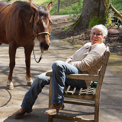 Karin Miller in sehr entspannter Haltung auf einem Stuhl- ihr Pferd steht entspannt neben ihr